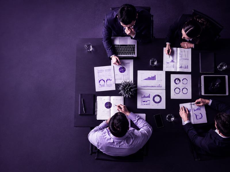 Vista aérea de uma reunião executiva, com profissionais reunidos ao redor de uma mesa analisando gráficos, relatórios e dados em notebooks.