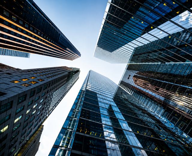 Perspectiva vertical entre edifícios corporativos contemporâneos, destacando arquitetura, escala e convergência de estruturas. A composição remete à visão estratégica, governança, solidez institucional e integração necessárias para estruturar a inovação n