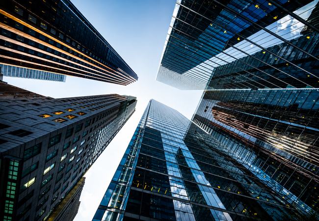Perspectiva vertical entre edifícios corporativos contemporâneos, destacando arquitetura, escala e convergência de estruturas. A composição remete à visão estratégica, governança, solidez institucional e integração necessárias para estruturar a inovação n