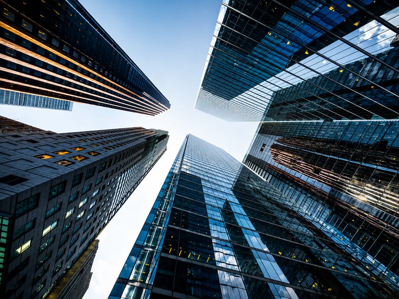Perspectiva vertical entre edifícios corporativos contemporâneos, destacando arquitetura, escala e convergência de estruturas. A composição remete à visão estratégica, governança, solidez institucional e integração necessárias para estruturar a inovação n