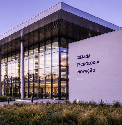 A imagem apresenta um edifício contemporâneo de pesquisa, ciência e inovação, com arquitetura institucional sofisticada, grandes fachadas de vidro, estrutura metálica e linhas geométricas precisas. O enquadramento em perspectiva valoriza a escala e a soli