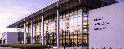 A imagem apresenta um edifício contemporâneo de pesquisa, ciência e inovação, com arquitetura institucional sofisticada, grandes fachadas de vidro, estrutura metálica e linhas geométricas precisas. O enquadramento em perspectiva valoriza a escala e a soli